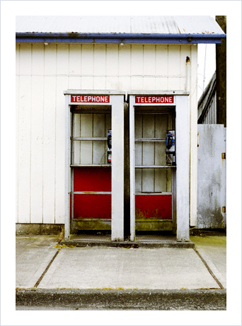 two phone booths on a sidewalk, standing against the side of a weathered white building